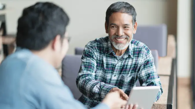 Happy old short beard asian man sitting, smiling and listen to partner that showing presentation on smart digital tablet.