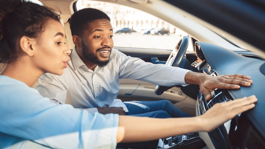 Afro couple at car showroom touching dash of new auto, sitting inside.
