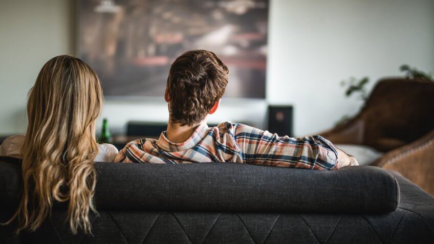 Rear view of a couple relaxing on sofa in the living room and watching a movie on TV.