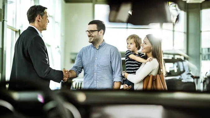 A happy family shakes a car salesman's hand while they stand in the car showroom.