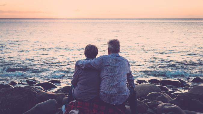 love and romance with adult  matures - elderly couple sitting and hugging each other looking at the sea at sunset relaxing.