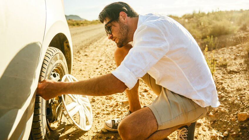 Shot of a young man inspecting a flat tyre on his car in a rural area.
