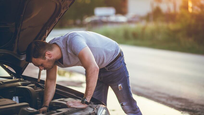 Young man is standing next to a broken down car, examining under the hood.