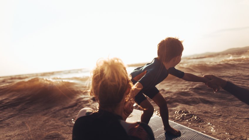 Photo of a cheerful little boy trying to stand up on a surfboard right before the wave strikes with a little help from his mother/ surfing teacher.