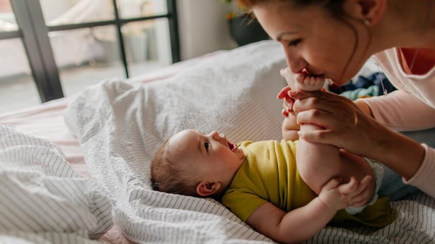 Portrait of a little smiling baby boy and his mom, kissing his tiny feet right after waking up in his nursery.