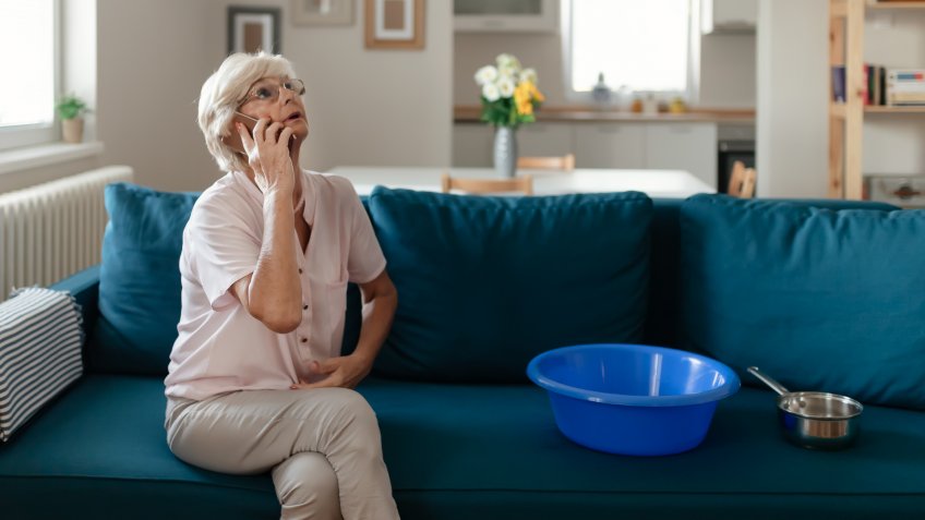 Shocked Senior Woman Calling Plumber While Collecting Water Leaking From Ceiling Using Utensil.