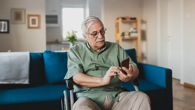 Handicapped senior person sitting in a wheelchair and using a smart phone at home in the living room.