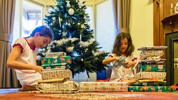Two smiling young sisters opening wrapped Xmas gifts with morning sun shining through window and Christmas tree in background.