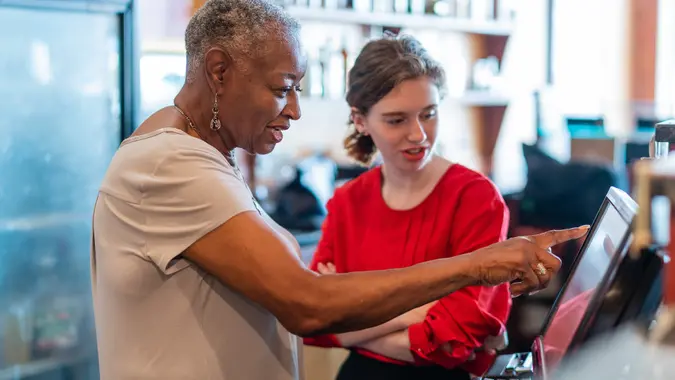 The active senior, 77-years-old, African-American businesswoman, business owner, teaching the new employee, the 18-years-old Caucasian white girl, how to use the computerized cash register in the small local restaurant.