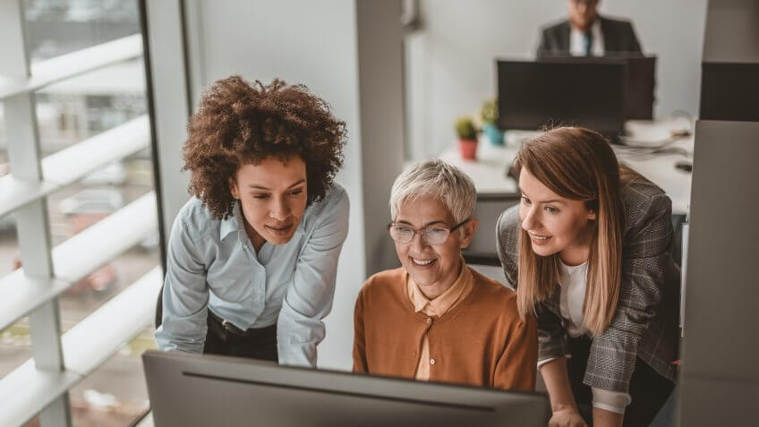 Photo of Mature businesswoman with female colleagues discussing at table in office while looking at desktop PC.