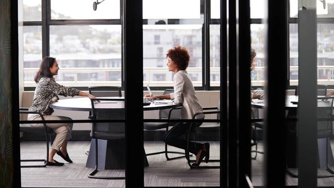 Two millennial businesswomen meeting for a job interview, full length, seen through glass wall.