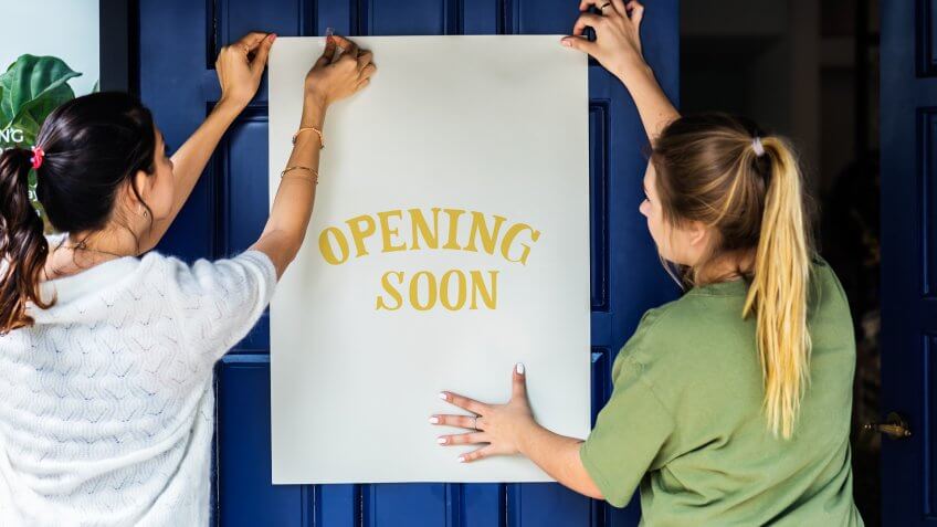 Women putting on store opening soon sign.