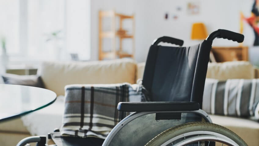 Empty wheelchair dor a disable person with folded plaid on background of big couch with cushions in living-room.