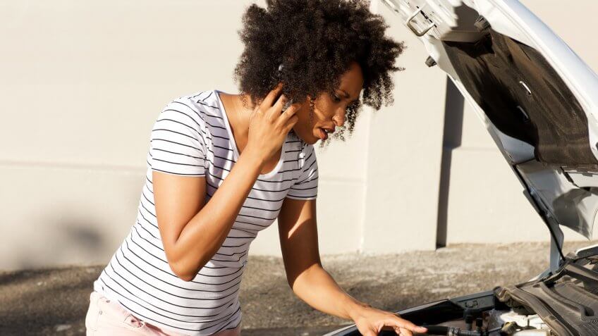 Portrait of young african woman standing by broken down car parked on the road and calling for assistance.