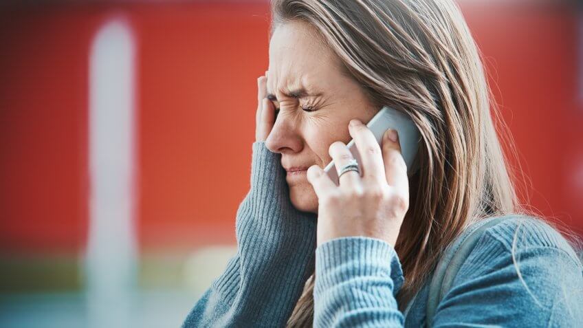 A young blonde woman grimaces, hand to her face as she hears something on her smart phone, obviously bad news.