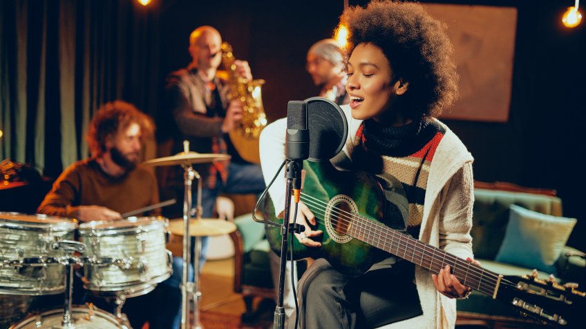 Mixed race woman singing and playing guitar while sitting on chair with legs crossed.