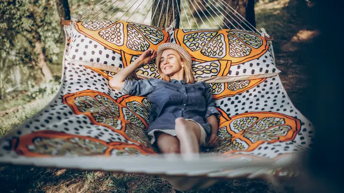 Smiling blonde hipster woman sleeping on a hammock outdoors, beautiful girl in stylish jean clothing relaxing on hammock in the park near a lake, camping concept.