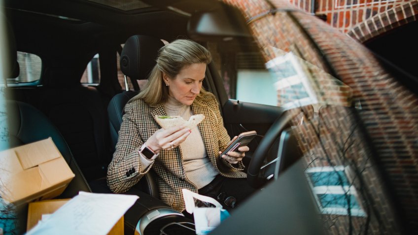 A side-view shot of a mature business woman sitting in the front seat of her car, she is using her mobile phone while she eats her lunch.