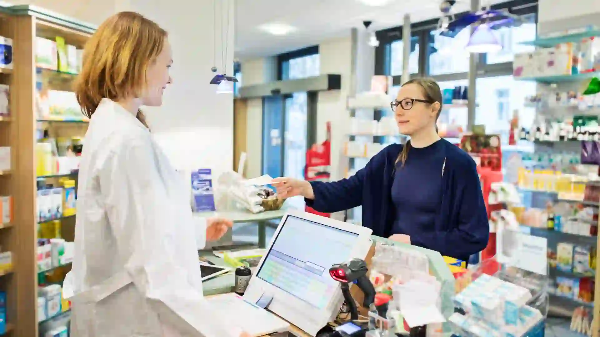 A woman hands a prescription to a female pharmacist behind the counter at a brightly lit pharmacy store.
