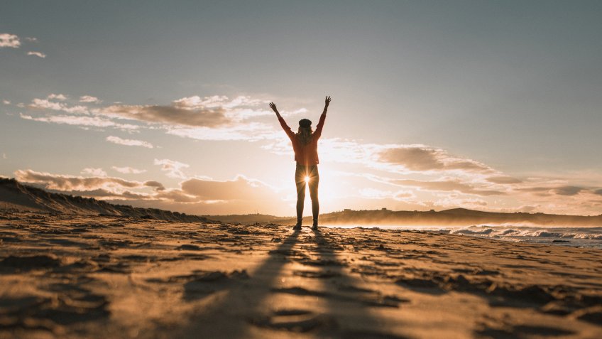 A young woman with arms raised at a beach at sunset.