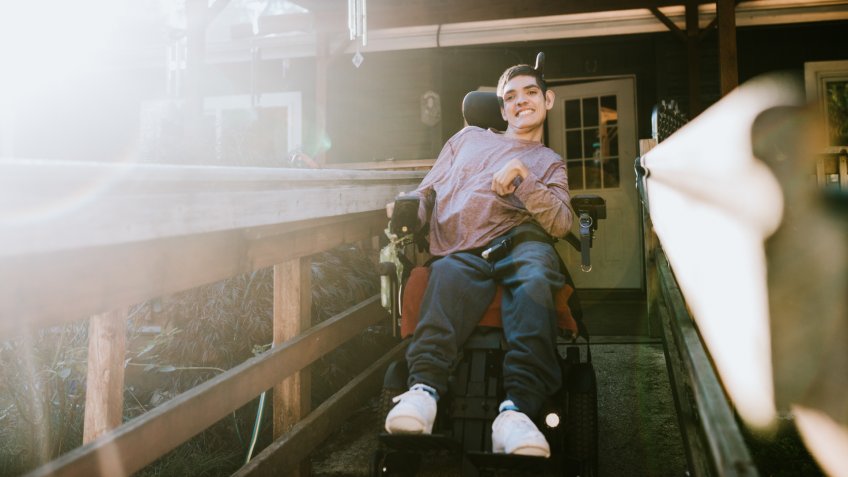 A cheerful young adult man with cerebral palsy drives down a wheelchair ramp to go outdoors on a bright sunny day.