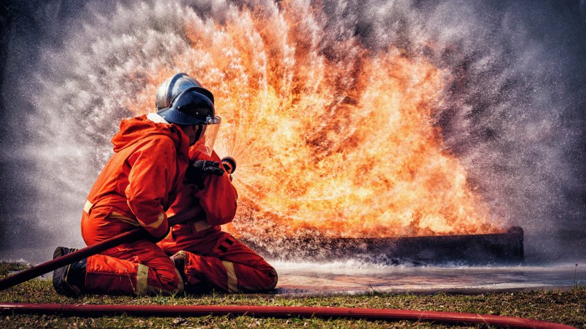 Firefighter in fire fighting suit spraying water, Firemen fighting raging fire with huge flames of burning, Fire prevention and extinguishing concept.