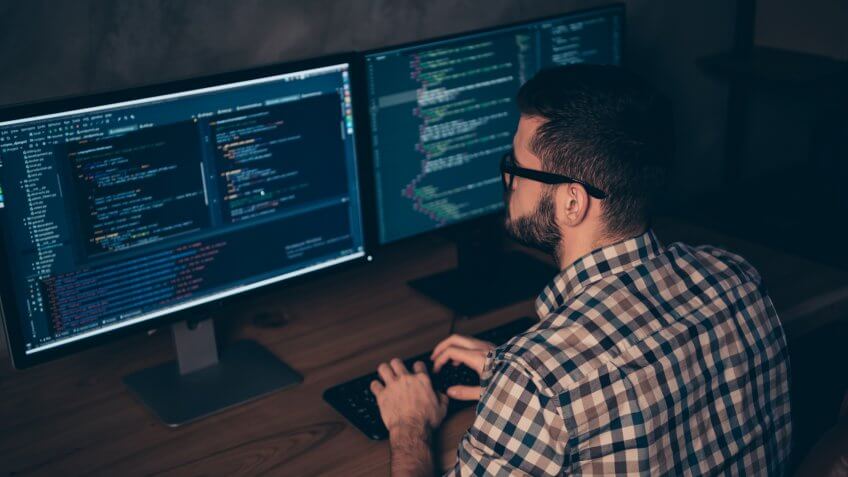 Close up back rear behind photo handsome he him his guy typing writing, keyboard development outsource IT processing language two monitors table office wear specs formalwear plaid shirt.