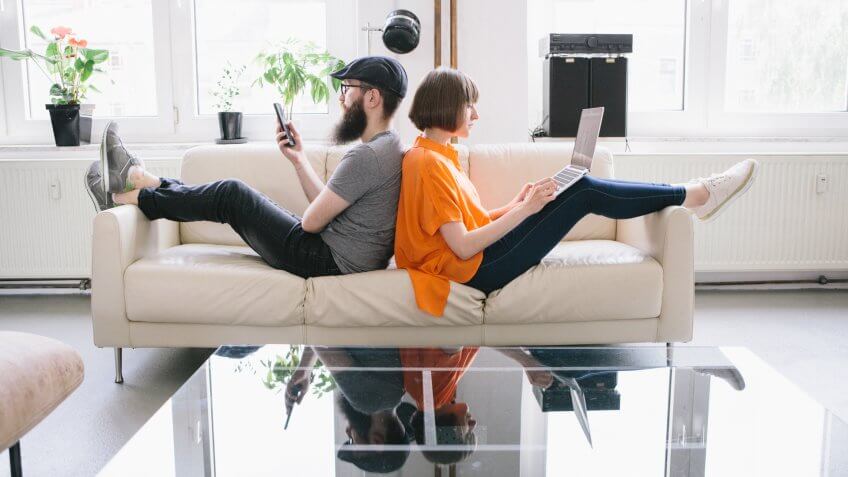 hipster couple sit back to back on couch and look into digital devices.