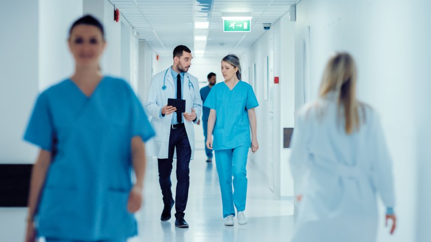 Surgeon and Female Doctor Walk Through Hospital Hallway, They Consult Digital Tablet Computer while Talking about Patient's Health.