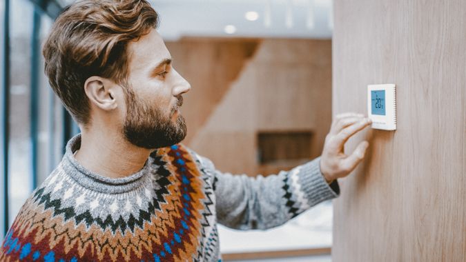Hombre con suéter sintiendo frío ajustando la temperatura ambiente con termostato electrónico en casa.