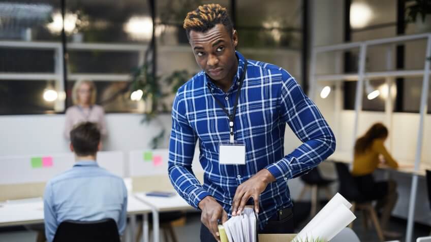 Serious frowning young black man with badge on neck discharged from place of employment checking paper files in box while leaving office.