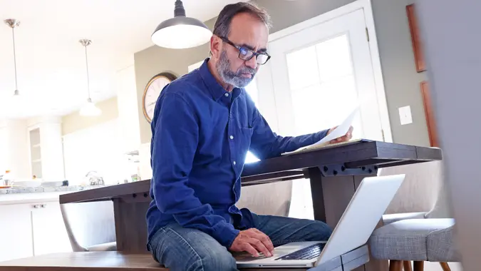 A Hispanic man in his late fifties sits on a bench at his dining room table and holds some paperwork in his hands as he works on his laptop computer.