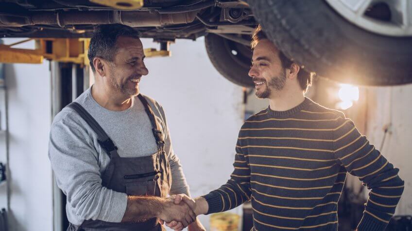 Mature adult auto mechanic, standing under the car, shaking hands with his customer, job is done.