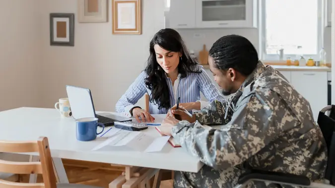 Disabled African American Male Soldier in a Wheelchair in Military Uniform With Female Financial Advisor Consulting About Financial Paperwork, Doing Household Accounting at Home.