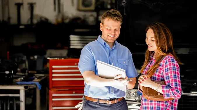Happy customer! A smiling Latin descent woman discusses automobile repair invoice with an auto mechanic in a repair shop.