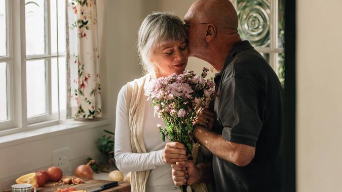 Senior couple standing in kitchen holding a bunch of flowers.