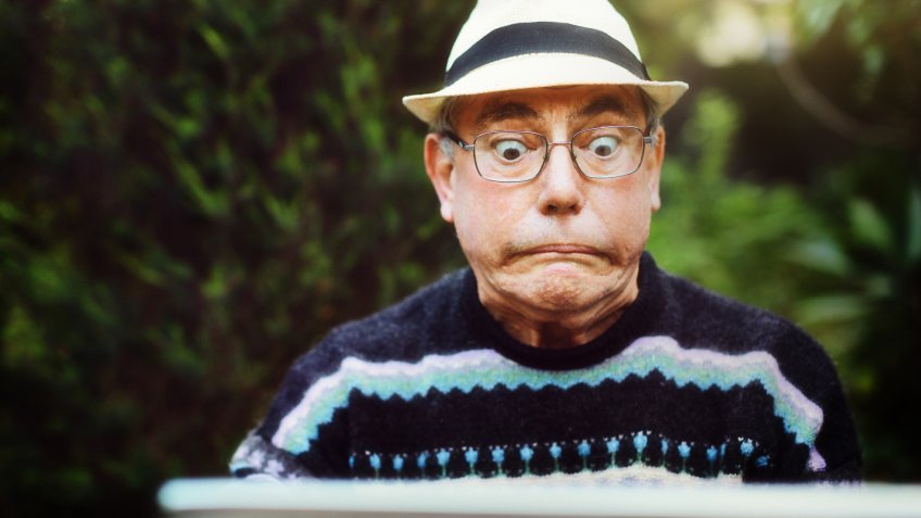 A senior man sitting in a garden using his laptop has a problem and glares at it, pulling a humorous face.