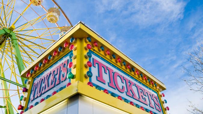 Tickets sign at county fair with Ferris wheel and light colored blue sky with white clouds in the background.