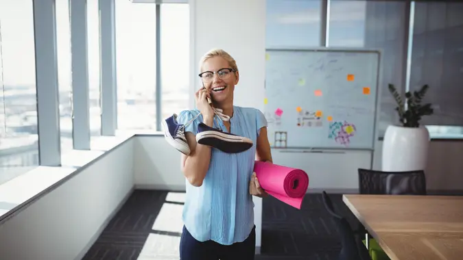 Smiling executive talking on mobile phone while holding exercise mat and shoes in office.