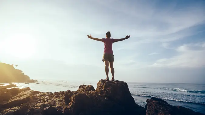 Freedom woman outstretched arms on sunrise seaside rock cliff edge.