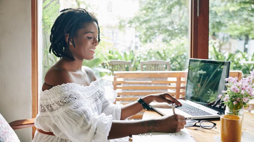 African happy woman sitting at the table in front of laptop and writing in document she working at home.