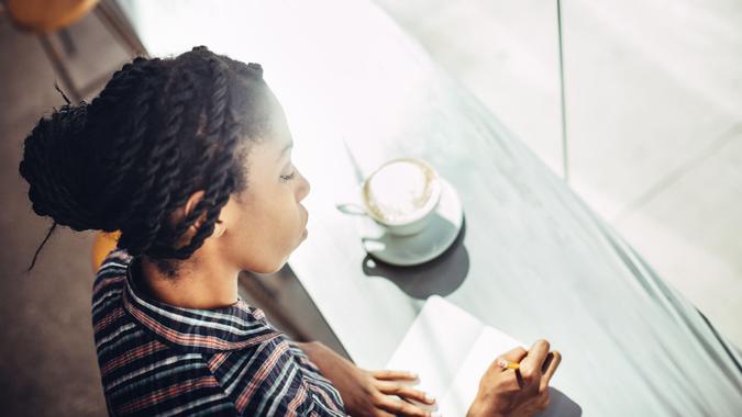 A beautiful young African American woman sits at the window counter of a coffee shop, enjoying a latte while writing ideas down in a small notepad journal or diary.