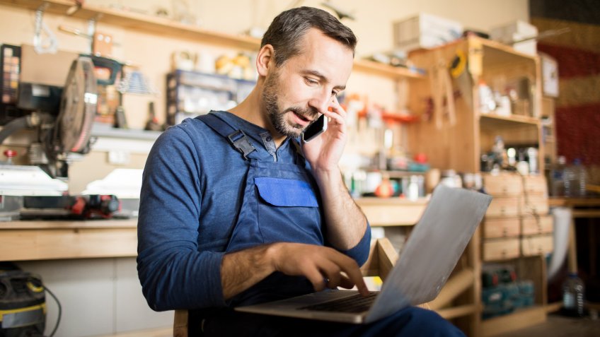 Portrait of mature worker using laptop and speaking by smartphone in workshop interior, copy space.