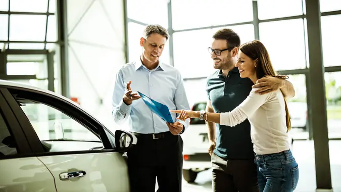 Happy embraced couple and car salesperson going through paperwork while buying a car in a showroom.