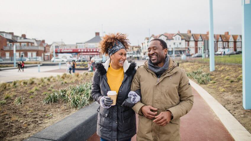 A couple walking arm-in-arm catching up over a coffee as they walk along a footpath.