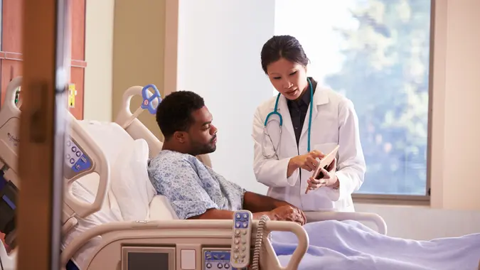 Hospital Doctor With Digital Tablet Talks To Male Patient.