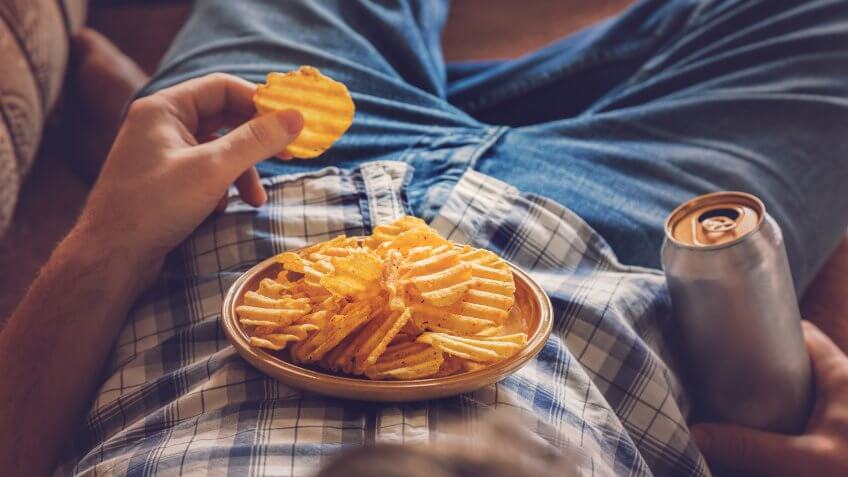 After work a guy wearing shirt and jeans lying on sofa, drinking a cold beer, eating crisps and watching sport tv channel.