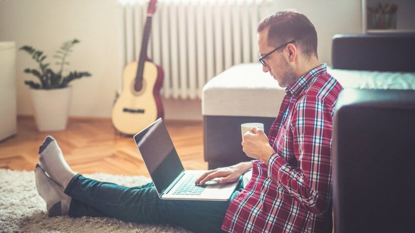 Young man sitting on the floor drinking coffee and surf the internet with lap top.
