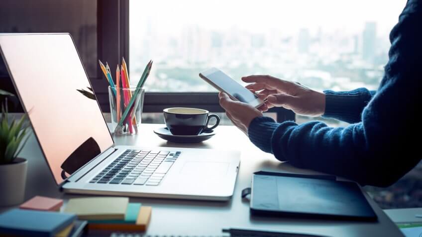 Young man using smartphone with modern work table and computer laptop and cityscapes view from window.