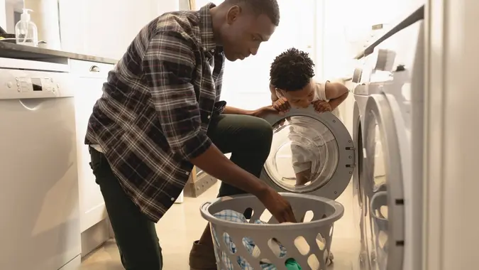 African American father and son washing clothes in washing machine at home.